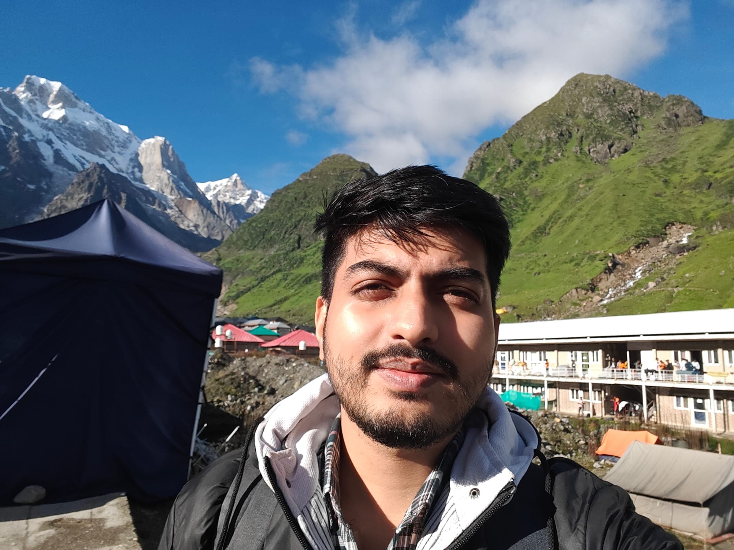 Biswajit in a black jacket, squinting in the sun, with snow peaks and green slopes behind him.