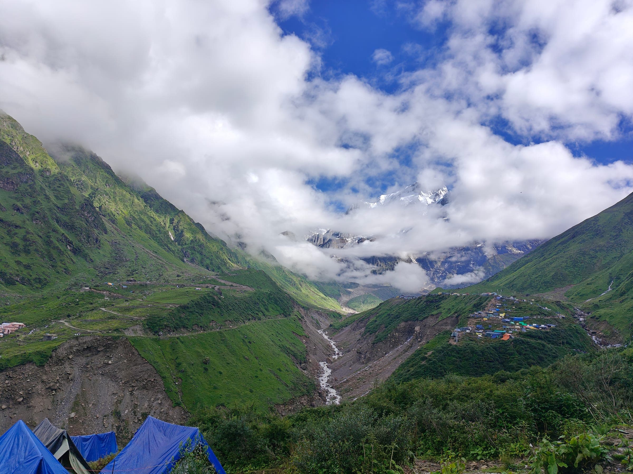 A glacial river running down a green valley towards a snow peak half hidden in cloud, blue tents in the foreground.